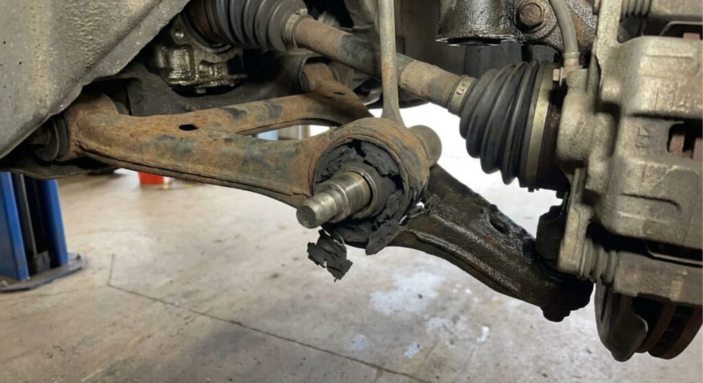 Vue sous un véhicule montrant un silent bloc en caoutchouc totalement déchiré et désolidarisé sur l'axe du triangle de suspension avant